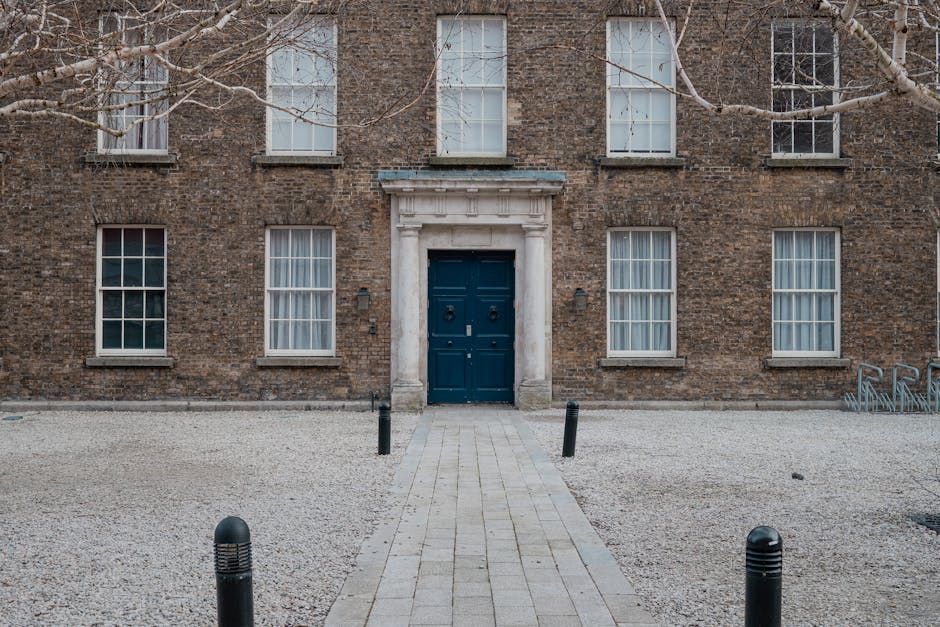 Exterior view of a brick building with six large sash windows, four on the ground floor and two on the upper floor, with white window frames and curtains. The centre features a dark blue wooden door set within a white stone surround, with a small columned portico above. The entrance area is paved with light-colored stone and bordered by a gravel surface. In front of the building, there is a narrow pathway lined with short black bollards. The scene is taken during daytime under overcast weather, with bare tree branches partially visible at the top left and right corners, suggesting a winter season. The image relates to home relocation and furniture transport, consistent with the services provided by Man with Van Bloomsbury, who may assist with loading or unloading in such premises during house moves or removals near Russell Square.