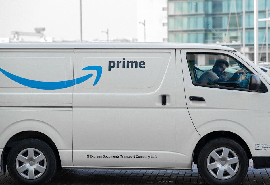 A white Amazon Prime delivery van parked outdoors on a paved area adjacent to a modern building with glass windows, where an individual inside the vehicle is seen using a mobile device. The van features the Amazon Prime logo with a blue arrow curved underneath the word 'prime' and the company name 'Q Express Documents Transport Company LLC' printed near the bottom. The vehicle is positioned close to a building entrance, suggesting possible loading or delivery activity related to home relocation or furniture transport. This scene captures the loading process or moving logistics, highlighting vehicle accessibility, the urban environment, and the use of packing materials inside the van area, relevant for house removals and packing and moving services by Man with Van Bloomsbury.