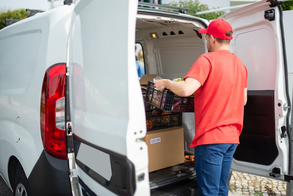 A man wearing a red cap and red t-shirt is loading groceries into the rear of a white delivery van parked outside a property. The inside of the van contains cardboard boxes and black plastic crates filled with various food items, some wrapped in plastic. He is placing a crate with fresh produce near other packages, with the van door open revealing the cargo space illuminated by interior lighting. The van is positioned on a cobblestone street, with a blurred background of greenery and a building. This scene depicts a typical home relocation or home delivery process, with careful handling of packaging materials and use of trolleys or straps for loading, consistent with professional removals and transport services offered by Man with Van Bloomsbury, supporting effective packing and furniture transport for house moves in the Bloomsbury area.