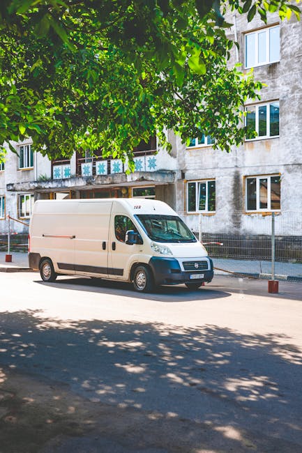 A white Amazon Prime delivery van parked outdoors on a paved area adjacent to a modern building with glass windows, where an individual inside the vehicle is seen using a mobile device. The van features the Amazon Prime logo with a blue arrow curved underneath the word 'prime' and the company name 'Q Express Documents Transport Company LLC' printed near the bottom. The vehicle is positioned close to a building entrance, suggesting possible loading or delivery activity related to home relocation or furniture transport. This scene captures the loading process or moving logistics, highlighting vehicle accessibility, the urban environment, and the use of packing materials inside the van area, relevant for house removals and packing and moving services by Man with Van Bloomsbury.