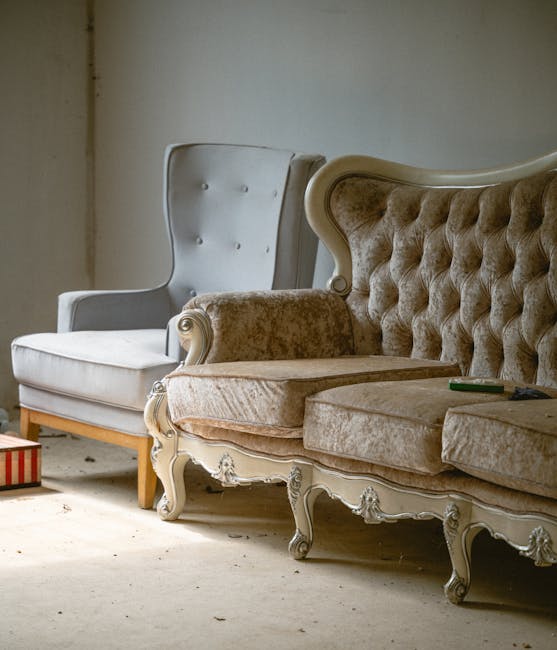 A collection of upholstered armchairs and a vintage-style sofa arranged indoors on a light-colored floor, with a plain wall in the background. The armchairs include a white fabric armchair with a high back and wooden legs, positioned to the left, and an ornate, cream-colored sofa with tufted upholstery, carved wooden details, and a beige velvet fabric, located on the right. The sofa appears to be partially disassembled for removal, with some cushions and fabric coverings loose or shifted. The environment suggests a home or storage space preparing for furniture transport. Visible materials include fabric, wood, and plastic wrap, and the lighting is soft and natural, indicating daytime. The scene reflects a furniture packing and loading process typical of house removals, with no people present but equipment such as a small box partially visible on the floor and the furniture arranged for eventual loading into a van, as managed by Man with Van Bloomsbury for home relocation or furniture transport services.