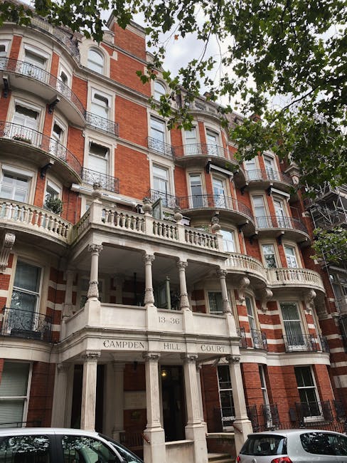The image shows the entrance of the Bloomsbury Street Hotel, a multi-storey building with a red brick exterior and white window frames. In front of the entrance, there is a black awning with the hotel's name, 'BLOOMSBURY STREET HOTEL,' displayed on it. A Union Jack flag is mounted on a pole attached to the building's facade, fluttering in the breeze. The entrance area is supported by beige stone pillars, and the doorway is partially visible beneath a white arch above the awning. The sidewalk in front is clean, with some greenery and small bushes along the sides. The scene is well-lit, suggesting it is daytime, and it captures the building's exterior, which could be relevant for house removals or moving logistics related to property upgrades or hotel relocations.