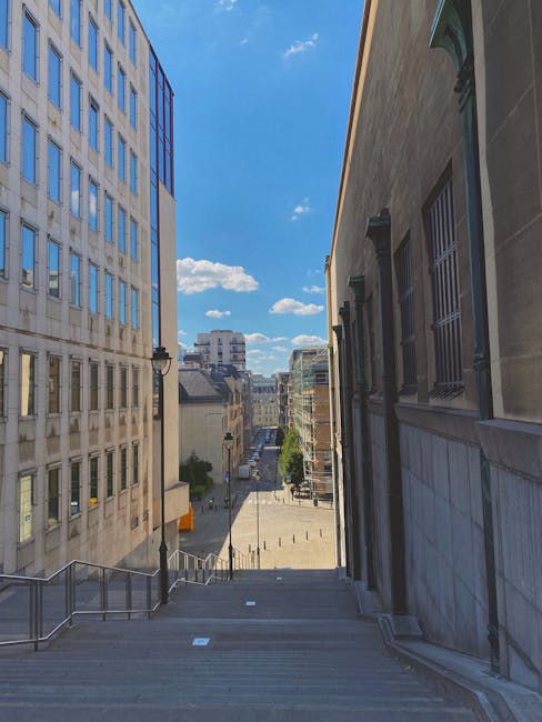 A photograph taken from the top of a steep exterior staircase outside a building, looking onto a narrow city street in Bloomsbury. The staircase has metal handrails on both sides, leading down to a paved area with short bollards and a few parked bicycles. To the left, there is a tall, modern office building with a grid of glass and concrete windows, while on the right, a historic stone building features barred windows and classical architectural details. The street below is lined with additional buildings, some with scaffolding and glass facades, under a bright blue sky with a few scattered clouds. This scene captures the urban environment typical of a home relocation or furniture transport process, with a focus on navigating narrow access points for removals services, as provided by Man with Van Bloomsbury.