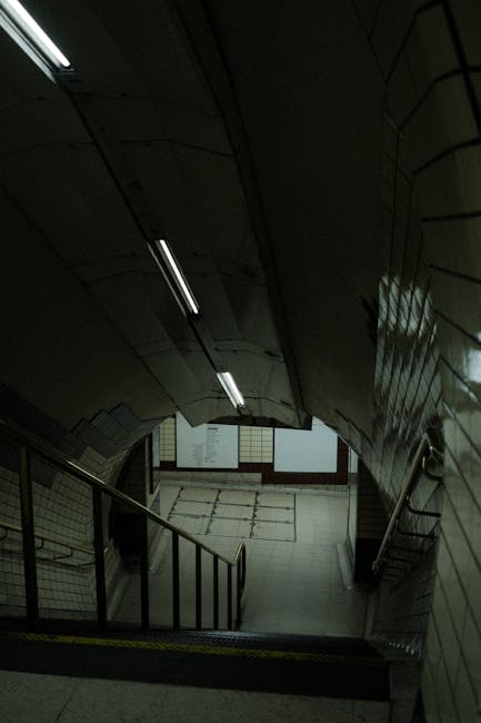 The image depicts a downward view of a staircase inside a building with metallic handrails and tiled walls, leading from an upper level to a lower landing illuminated by fluorescent lighting. The staircase appears narrow, with a slight curve, and is part of an indoor passageway likely within a residential or commercial property. The lower area features a tiled floor and a partially open door at the bottom. This scene may represent an interior space involved in a house relocation or moving process, as managed by Man with Van Bloomsbury, who specialise in removals and furniture transport. The environment is utilitarian and functional, suitable for loading or unloading belongings during a home or apartment move, particularly in buildings with restricted access or narrow stairways, such as in historic or apartment buildings in the Bloomsbury area.