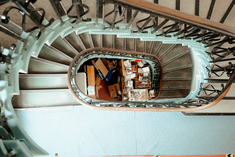 A photograph taken from the top of a steep exterior staircase outside a building, looking onto a narrow city street in Bloomsbury. The staircase has metal handrails on both sides, leading down to a paved area with short bollards and a few parked bicycles. To the left, there is a tall, modern office building with a grid of glass and concrete windows, while on the right, a historic stone building features barred windows and classical architectural details. The street below is lined with additional buildings, some with scaffolding and glass facades, under a bright blue sky with a few scattered clouds. This scene captures the urban environment typical of a home relocation or furniture transport process, with a focus on navigating narrow access points for removals services, as provided by Man with Van Bloomsbury.