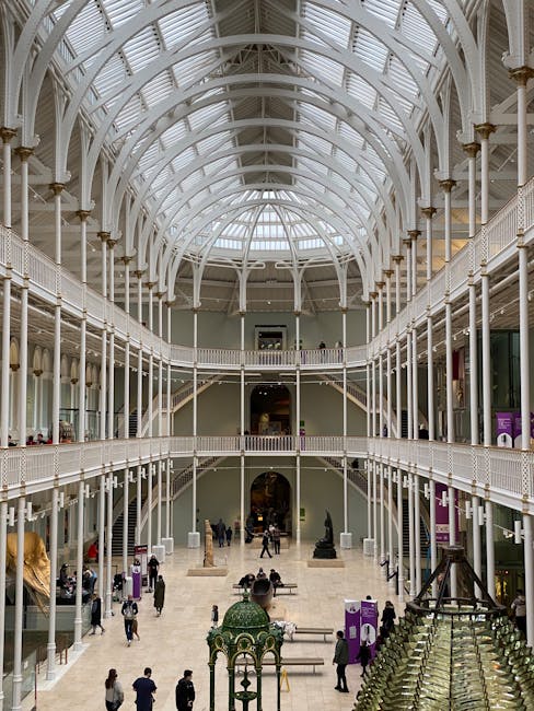 The image depicts the expansive interior of the British Museum, featuring a large central courtyard with multiple levels of white-painted ironwork balconies and railings. The high, arched glass roof allows natural light to illuminate the space, highlighting the classical architectural details. On the ground floor, visitors are seen walking, standing, and observing exhibits, while some are seated on benches near sculptures and display cases. The upper balconies are accessible via staircases, and some visitors are visible on the walkways overlooking the central area. Large sculptures and display objects are positioned at the far end of the courtyard, with informational signage and purple banners visible throughout the space. The scene captures the atmosphere of a busy cultural institution, with a focus on the architectural beauty and the movement of people within the museum, relevant to themes of interior space layout and visitor activity during a house or museum move or refurbishment, through the context of professional removals and transportation.