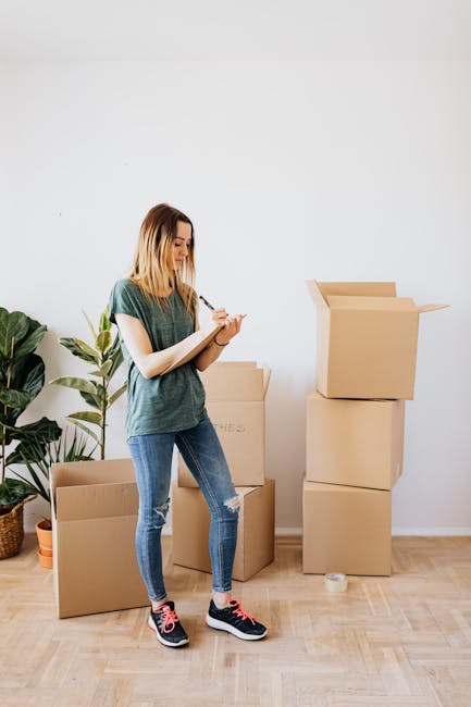 A young woman with shoulder-length blonde hair, dressed in a green t-shirt, ripped blue jeans, and black and pink sneakers, stands inside a room with light wooden flooring and a plain white wall. She is holding a pen and a notepad, appearing to take notes. Around her are several cardboard moving boxes, some stacked, with one open top box on the right side showing packing materials inside. To the left, there is a large potted plant with broad green leaves, adding a touch of greenery to the space. The scene depicts a packing or home relocation process, relevant to house removals and moving services such as those offered by Man with Van Bloomsbury, as part of the preparation for moving out of a WC1 studio in the Gower Street area.