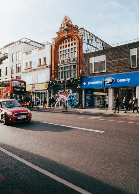 View of a busy street in Camden with a row of commercial buildings, including a pharmacy and beauty shop, and pedestrians walking along the pavement. A red double-decker bus and a car are visible on t