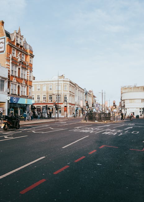 View of a busy street in Camden with a row of commercial buildings, including a pharmacy and beauty shop, and pedestrians walking along the pavement. A red double-decker bus and a car are visible on t