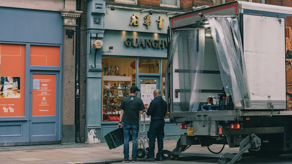 Two movers, dressed in dark clothing, stand outside a storefront with signage featuring Chinese characters and the word 'GUANG' on a blue facade. One mover carries a small cardboard box, while the other pushes a hand trolley loaded with similar packages. A large, open-sided moving truck with a transparent plastic covering on one side is parked on the pavement, adjacent to the storefront. Inside the truck, neatly stacked boxes and packing materials are visible. The scene is set during daylight, with the building's architecture and street environment clearly visible, illustrating the process of home relocation and furniture transport, with attention to packing and loading procedures as part of a professional removals service provided by Man with Van Bloomsbury.