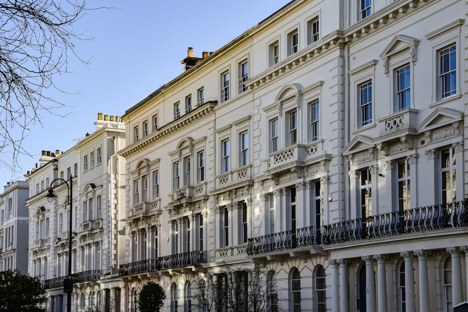 Exterior view of a row of classic Victorian-style terraced houses with white facades, ornate architectural details, and black wrought-iron balconies. The buildings are multi-storey with large sash windows, decorative mouldings, and cornices, illuminated by natural daylight. A black street lamp is visible on the pavement in front of the houses, with leafless trees partially visible on the left side of the image. The scene depicts a typical urban residential area suitable for house removals and furniture transport, emphasizing the importance of careful packing and loading processes facilitated by professional moving services such as those offered by Man with Van Bloomsbury. The photograph captures the curb appeal and structural elements of the properties, providing context for home relocation or moving logistics within a historic London neighbourhood.